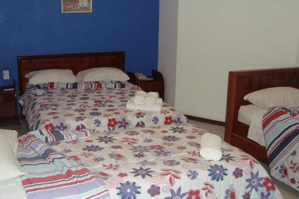 Una habitación con dos camas, cada una con una colcha de motivos florales y almohadas blancas. Se observa un cabecero de madera, una mesita de noche y una pequeña mesa con una silla.