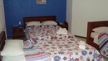 A room with two beds, each with a floral-patterned bedspread and white pillows. There is a wooden headboard, a nightstand, and a small table with a chair.