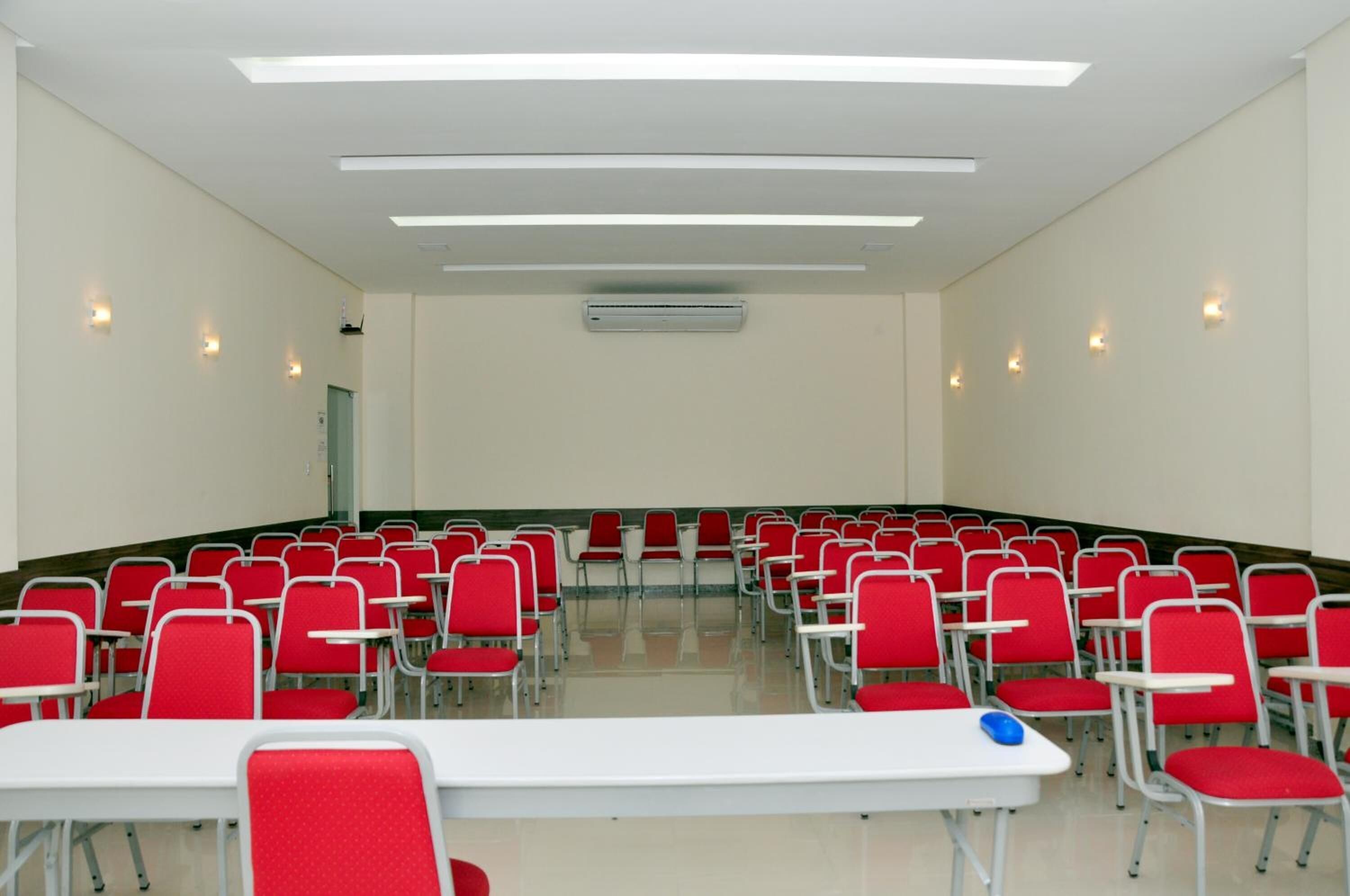 Une salle de conférence avec des rangées de chaises rouges et des tables blanches, équipée de luminaires au plafond et d’un système de climatisation.