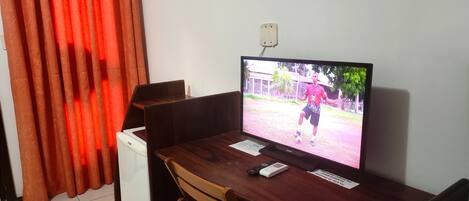 A room with a wooden desk, a television displaying a soccer player, a white appliance, and a red curtain.