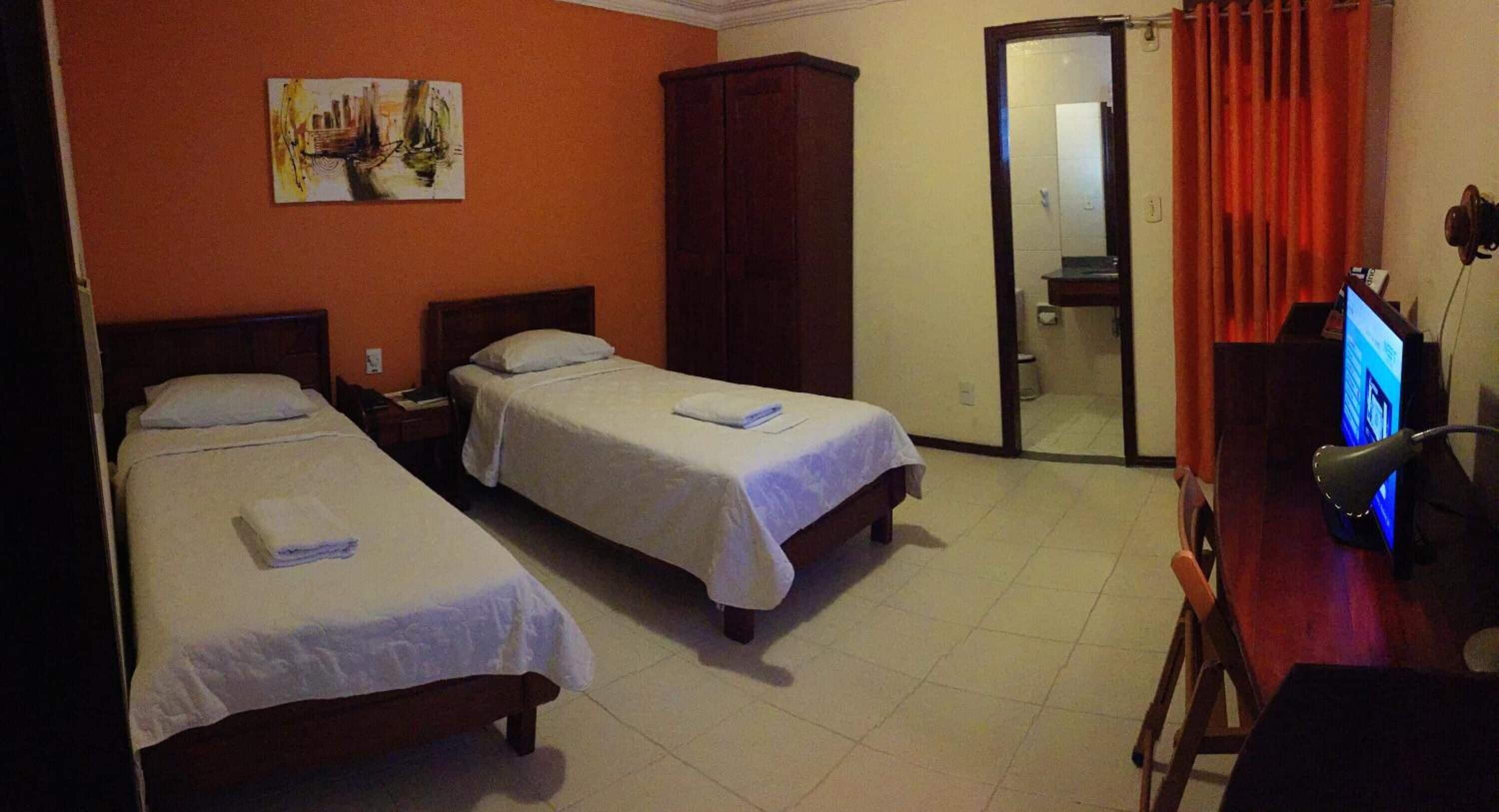Ein Hotelzimmer mit zwei Einzelbetten, einem Kleiderschrank, einem Schreibtisch mit Computer und einem durch eine offene Tür einsehbarer Badezimmer.