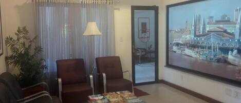 A hotel lobby with two leather armchairs, a coffee table with magazines, a large window with a cityscape view, and an air conditioning unit.