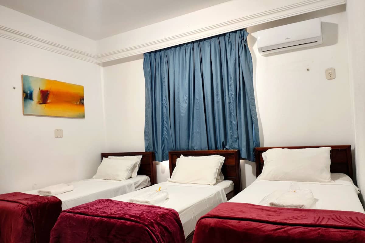 Habitación de hotel con tres camas individuales, cada una con colcha roja y almohadas blancas. Se observa una cortina azul, un cuadro en la pared y un equipo de aire acondicionado sobre las camas.