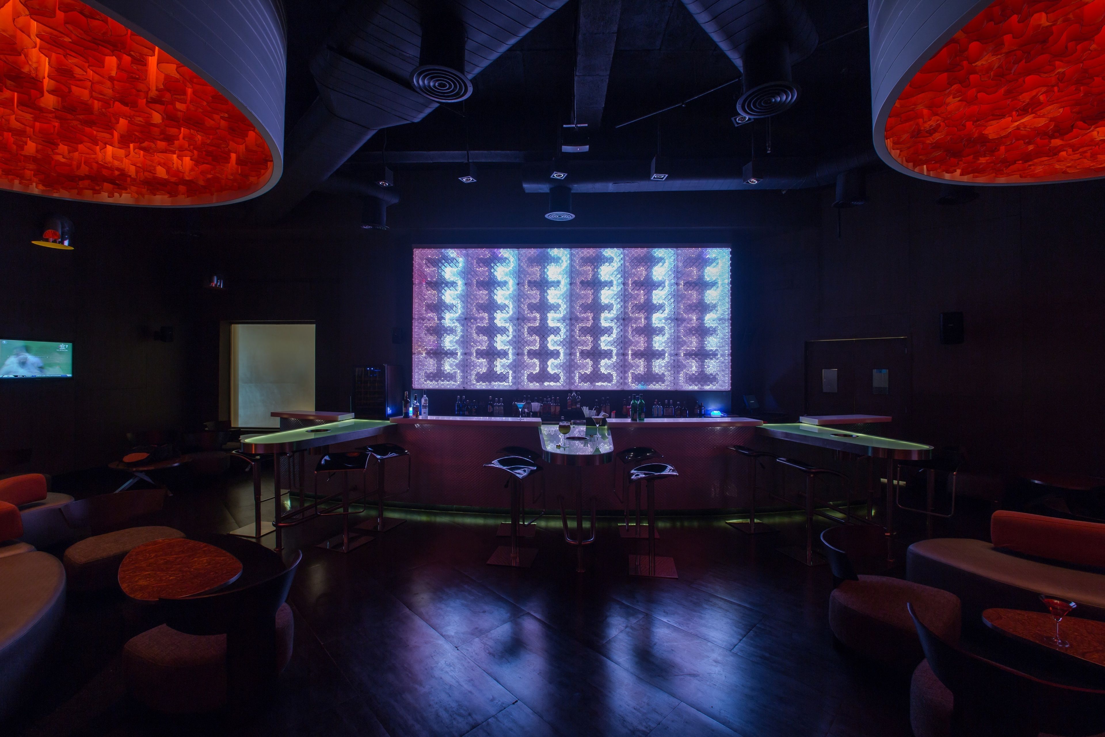 A dimly lit bar area with a large screen displaying a pattern, bar stools, and a counter with bottles.