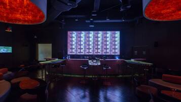 A dimly lit bar area with a large screen displaying a pattern, bar stools, and a counter with bottles.