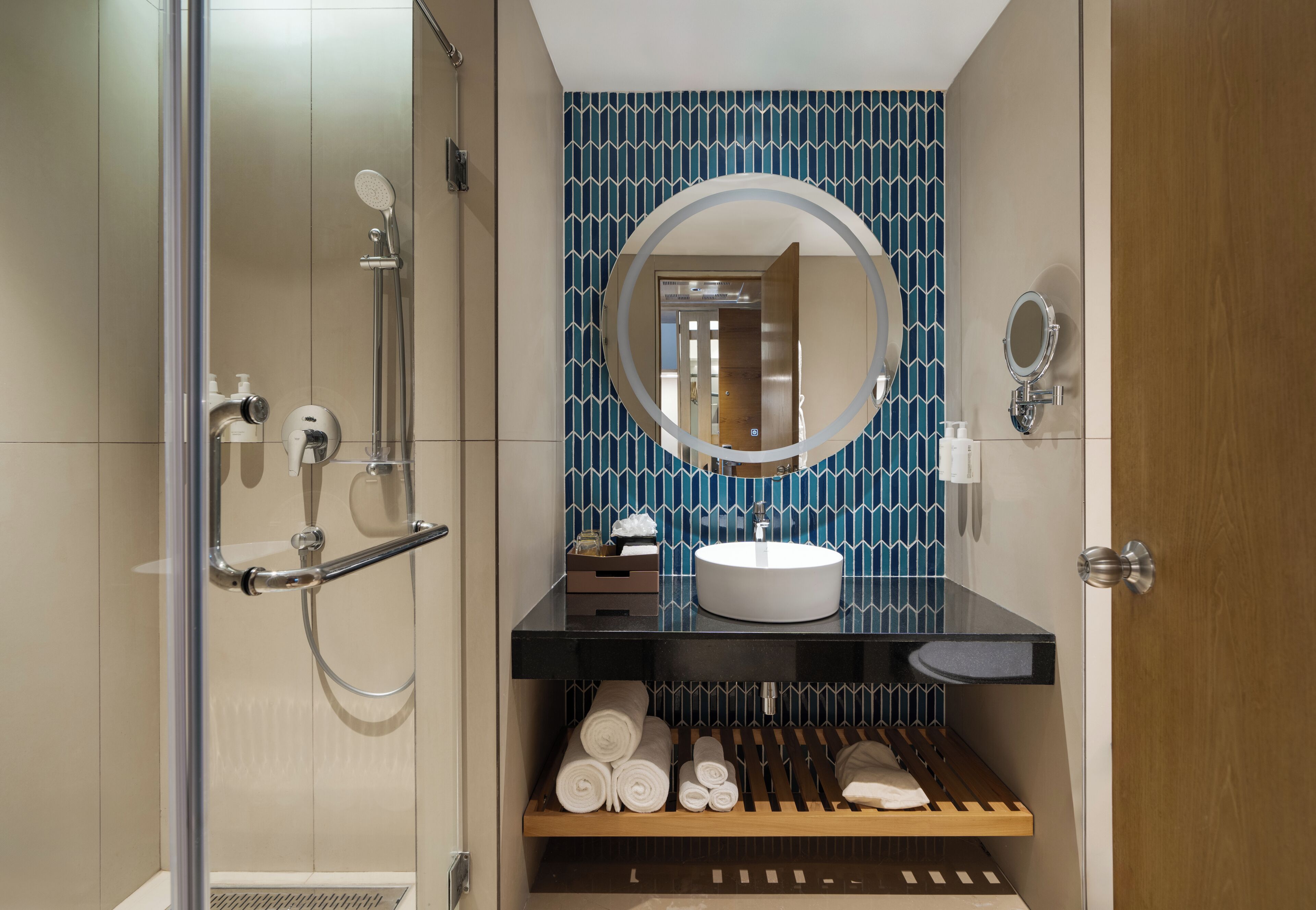 A modern bathroom with a glass shower enclosure, a round mirror, a white sink, and neatly rolled towels on a shelf.