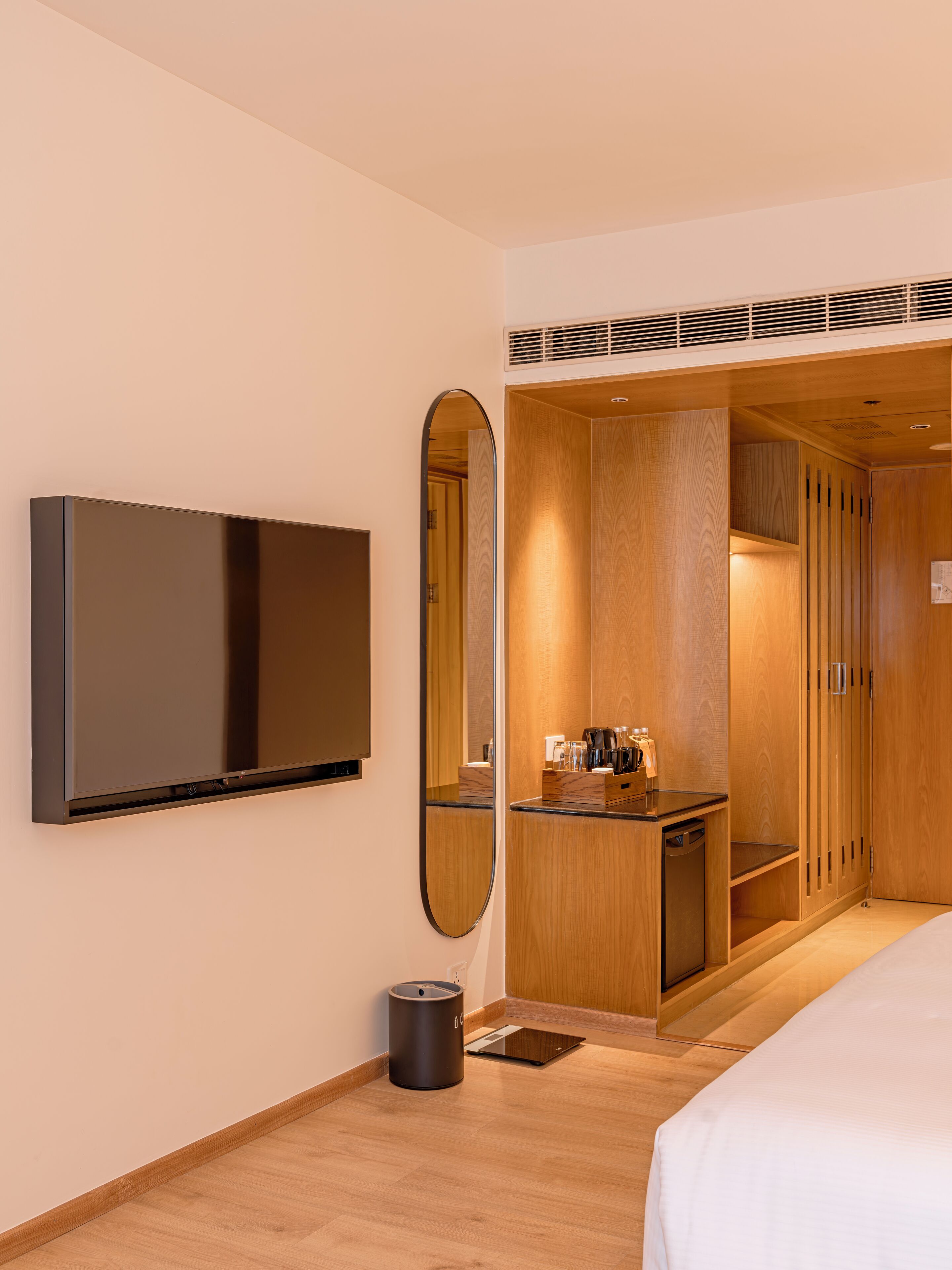 A hotel room with a flat-screen TV mounted on the wall, a wooden wardrobe, a small desk with a mirror, and a bed.