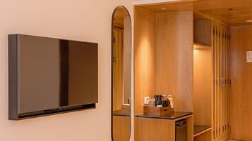A hotel room with a flat-screen TV mounted on the wall, a wooden wardrobe, a small desk with a mirror, and a bed.