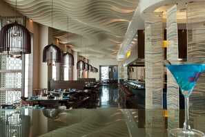 A modern restaurant interior with a wavy ceiling design, hanging pendant lights, and a bar area.