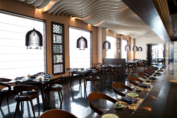 A modern restaurant with long communal tables, black chairs, and hanging pendant lights.