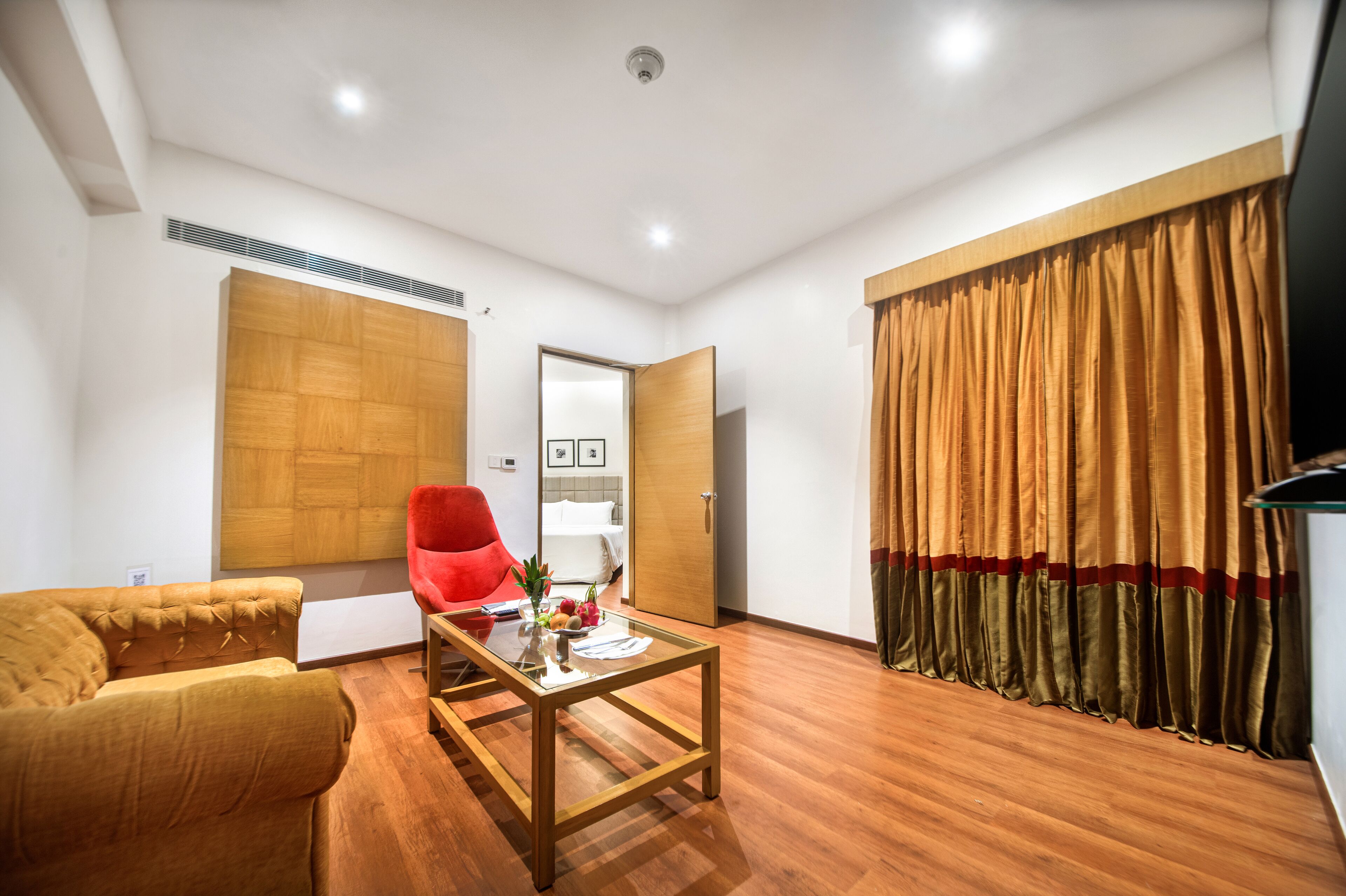 A modern hotel room with a wooden floor, a red armchair, a glass coffee table, a wardrobe, and a large window with curtains.