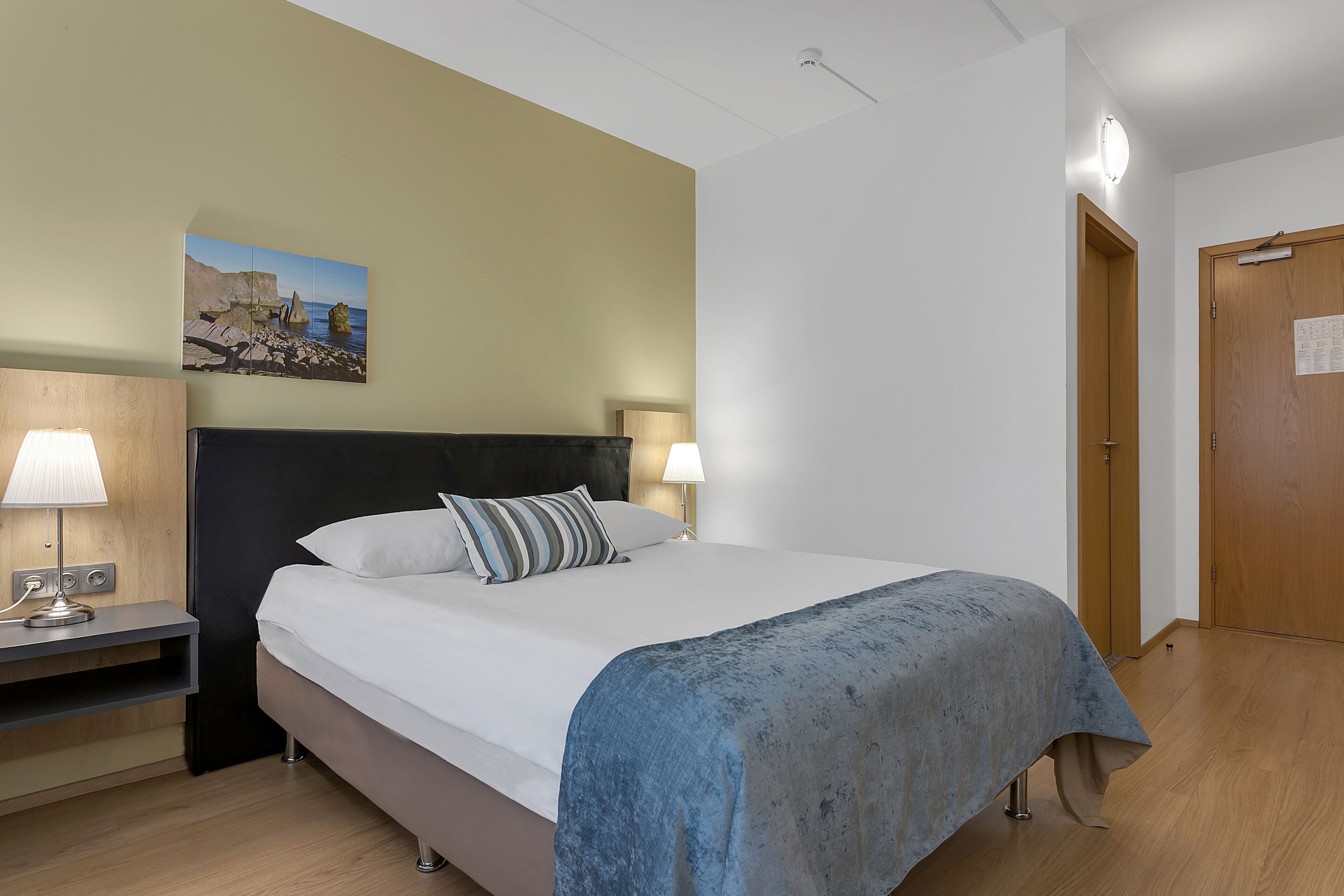 Habitación de hotel con cama, lámparas de noche, mesita de noche, puerta de madera y un cuadro colgado en la pared.