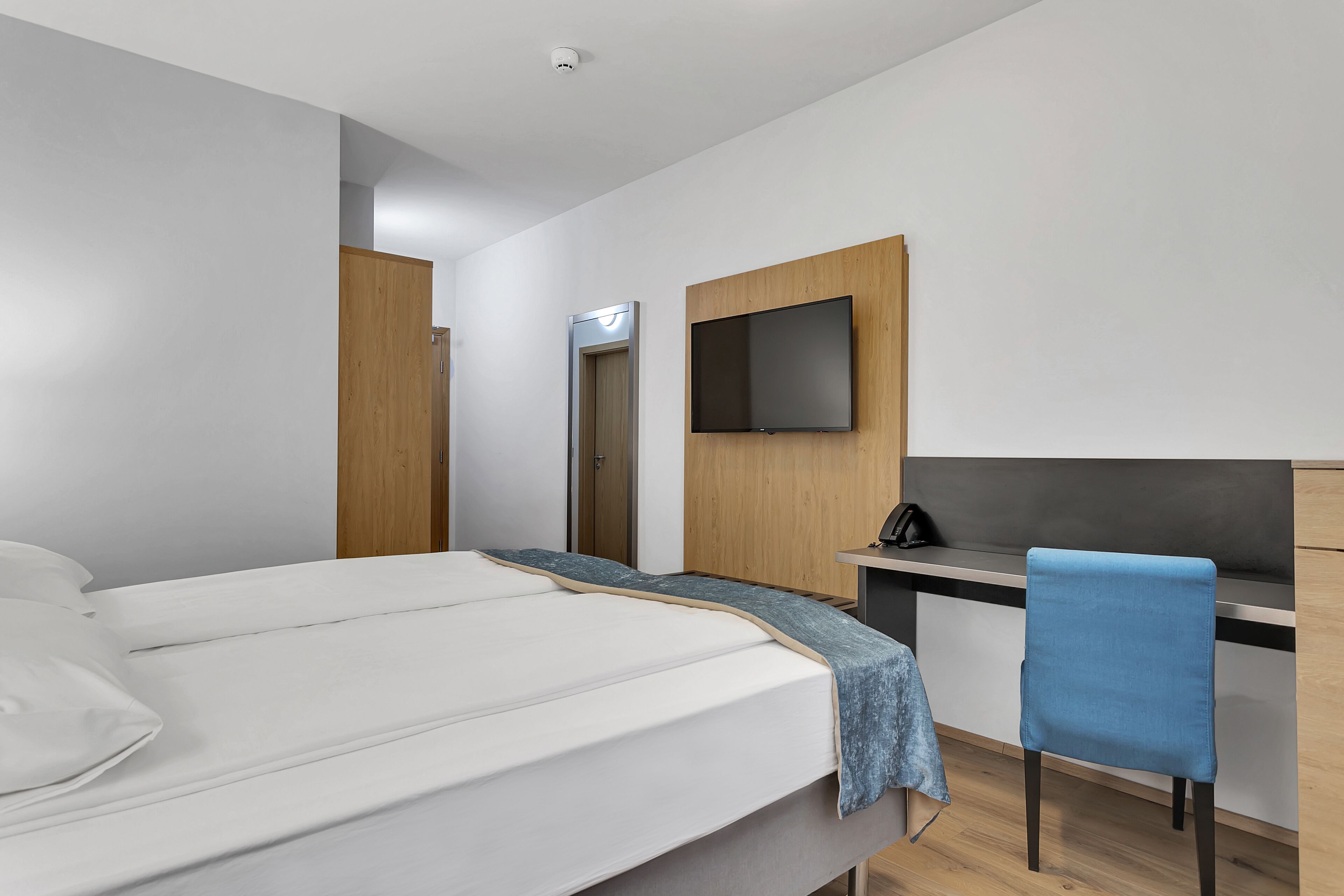 A modern hotel room with a bed, a desk, a chair, a TV mounted on the wall, and a door leading to another room.