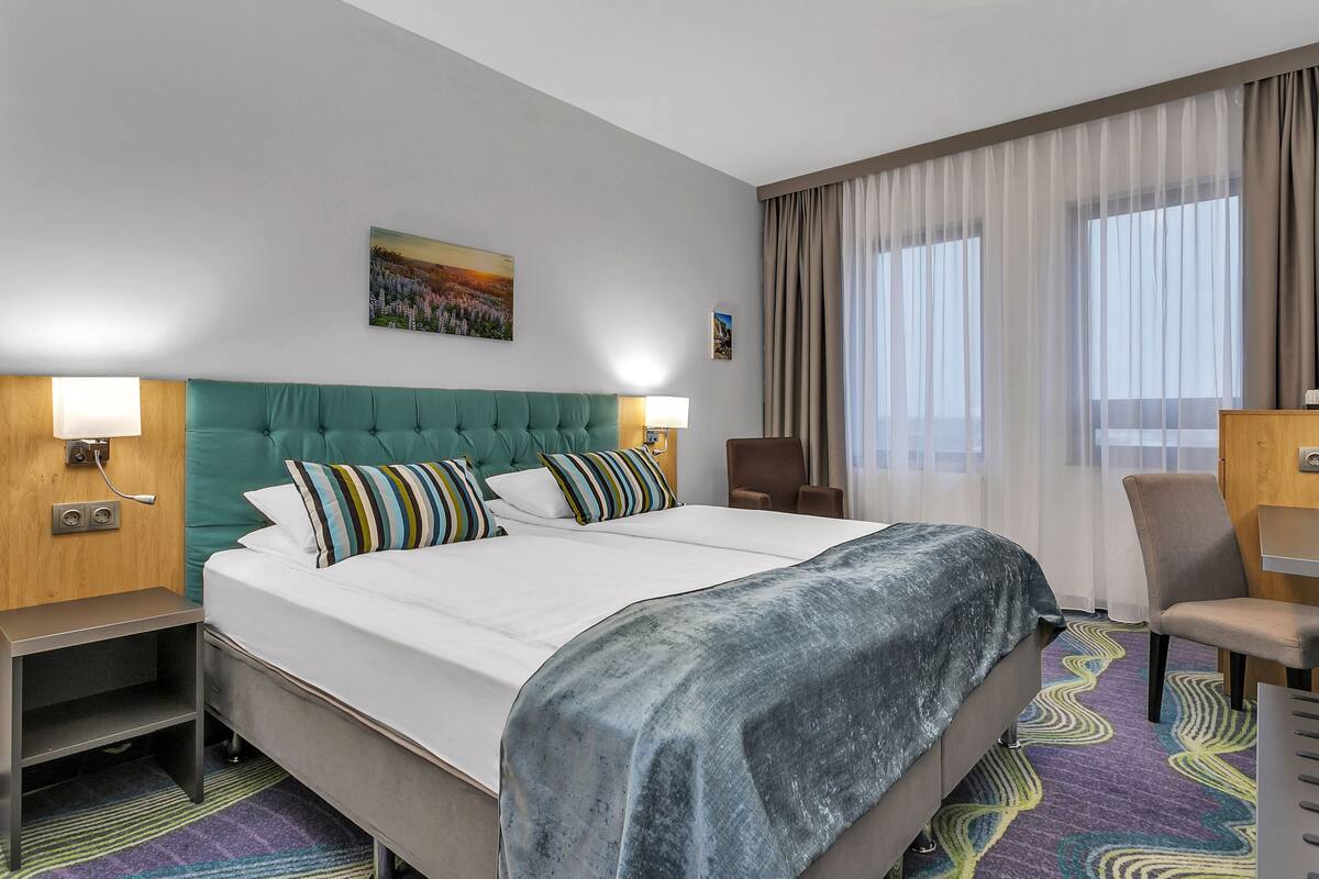 Habitación de hotel con una cama grande, un cabecero verde, dos mesitas de noche con lámparas, una silla y una ventana con cortinas traslúcidas.