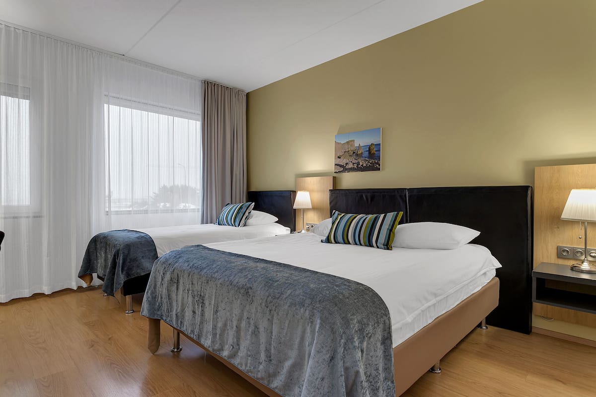 Habitación de hotel con dos camas, un ventanal con cortinas translúcidas, un cuadro en la pared y una mesita de noche con lámpara.