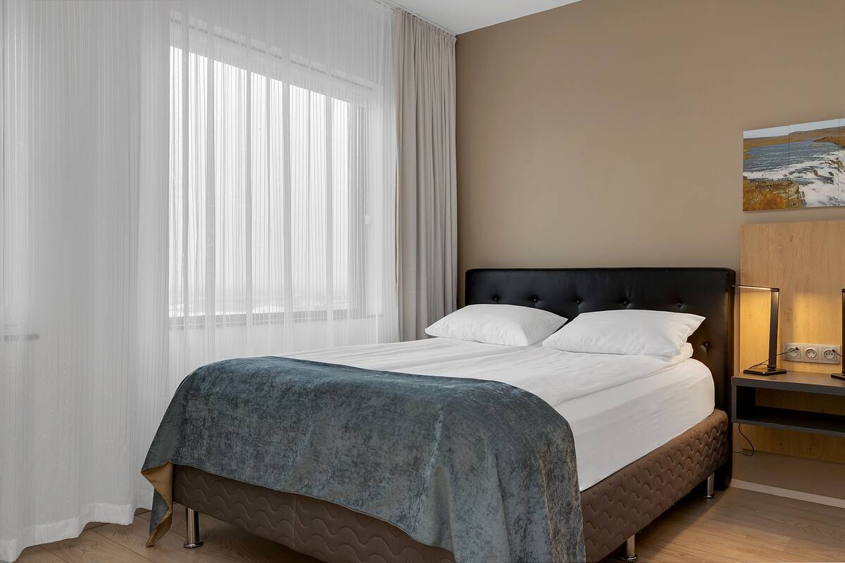 Una cama bien hecha con un cabecero oscuro, una mesita de noche con lámpara y un cuadro en la pared.