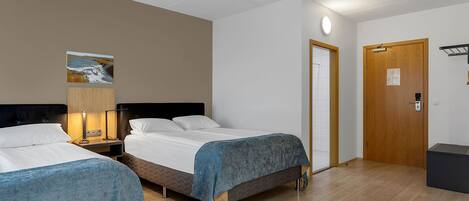 A hotel room with two beds, a wooden door, a small table with a lamp, and a painting on the wall.