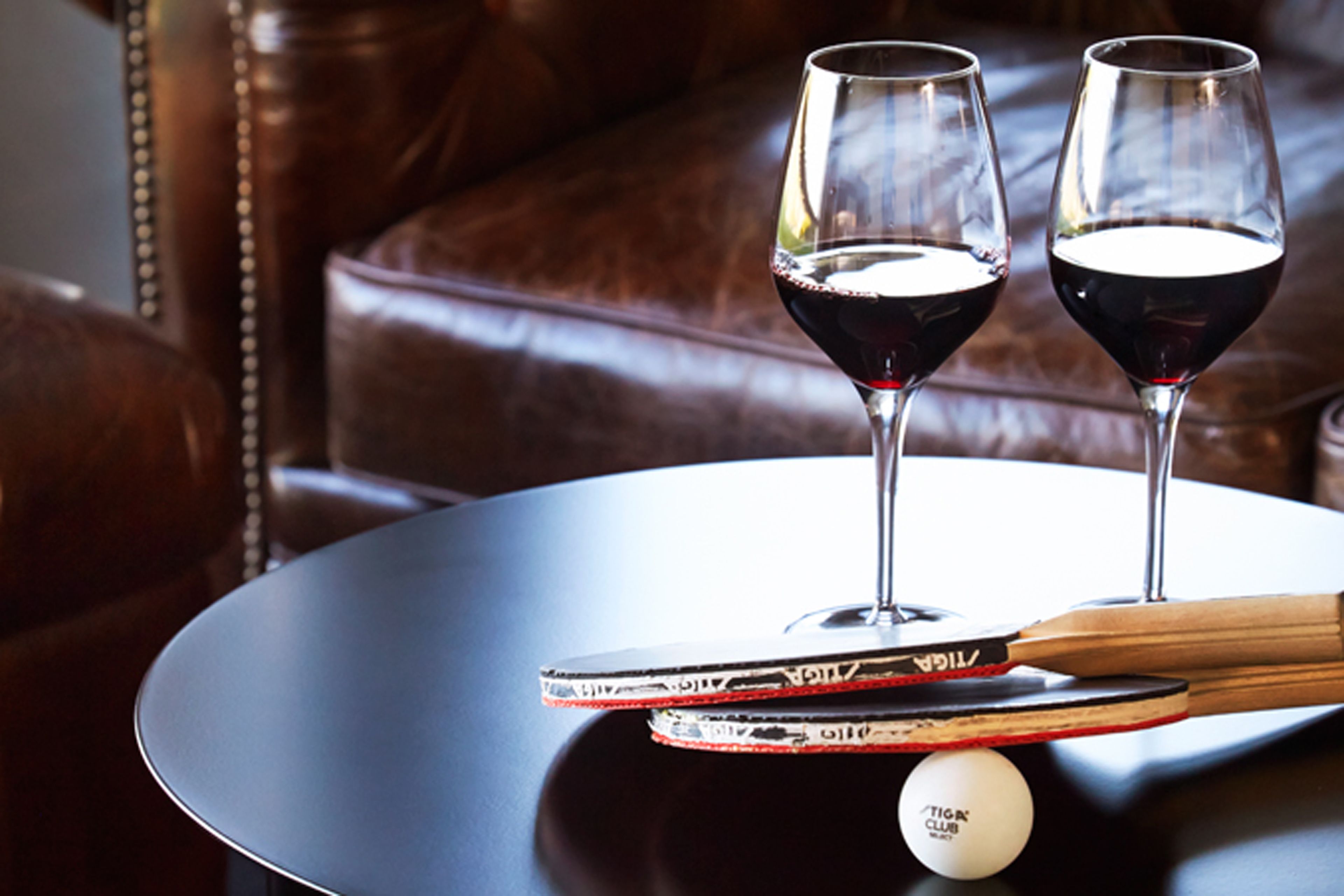 Dos copas de vino sobre una mesa redonda, junto con una paleta de ping pong y una bola blanca.