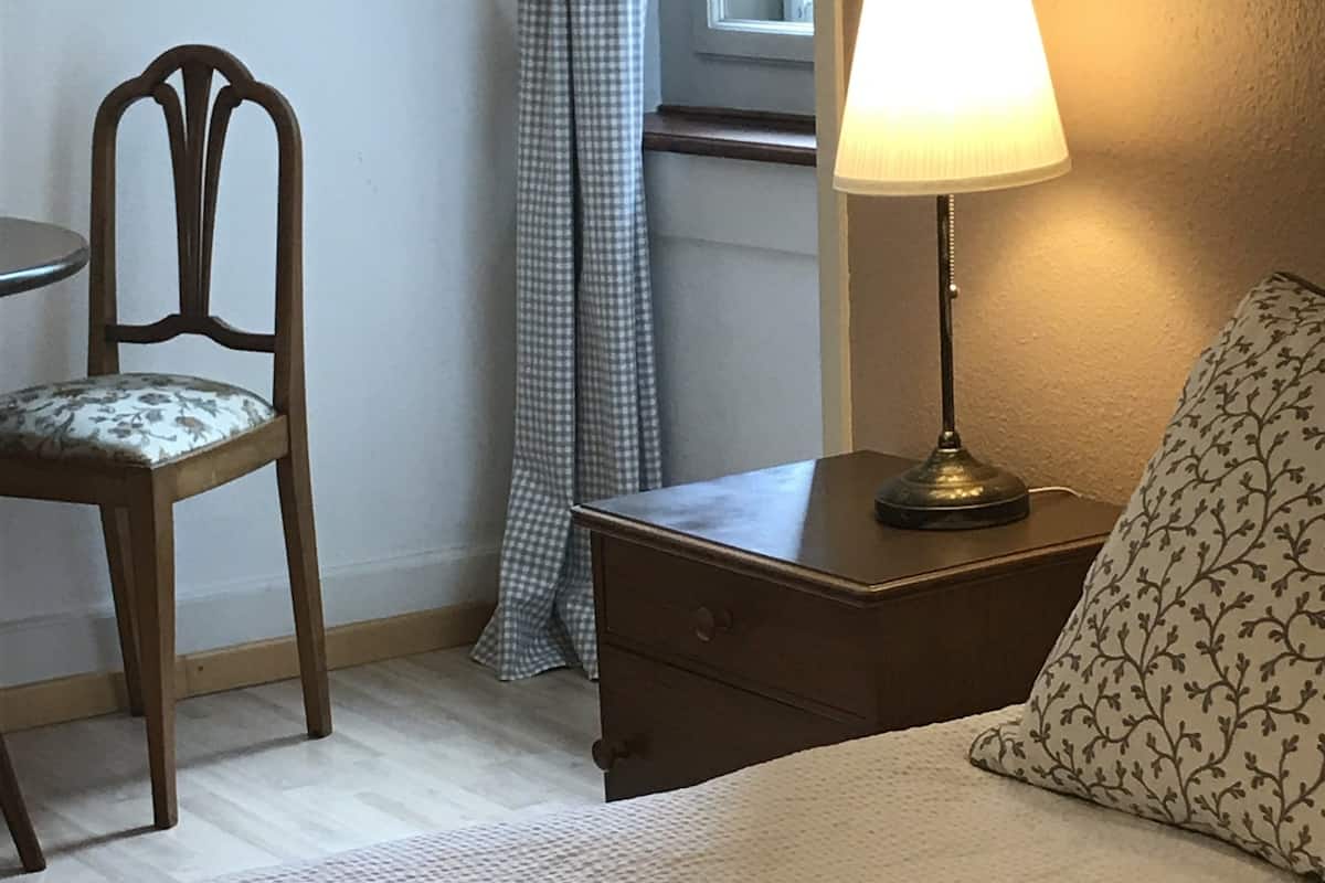 A neatly arranged bedroom with a bed, a chair, a bedside table with a lamp, and a window with curtains.