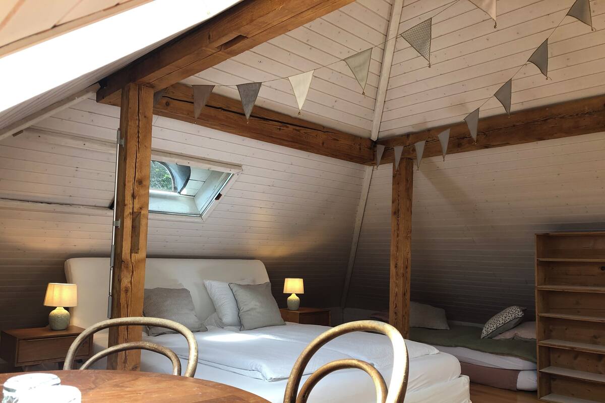A cozy attic bedroom with a wooden ceiling, a skylight, a bed with pillows, a bedside table with a lamp, and a wooden chair.