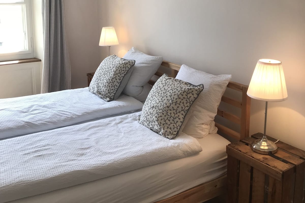 A double bed with two pillows, a bedside table with a lamp, and a window with curtains.