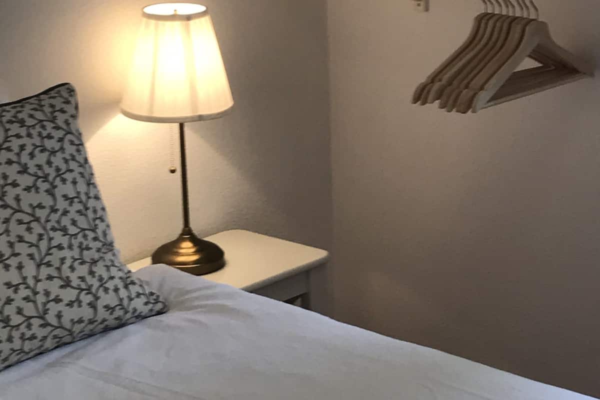 A neatly made bed with a white comforter and a patterned pillow, a bedside lamp, a small white nightstand, a potted plant, and a clothing rack with hangers.