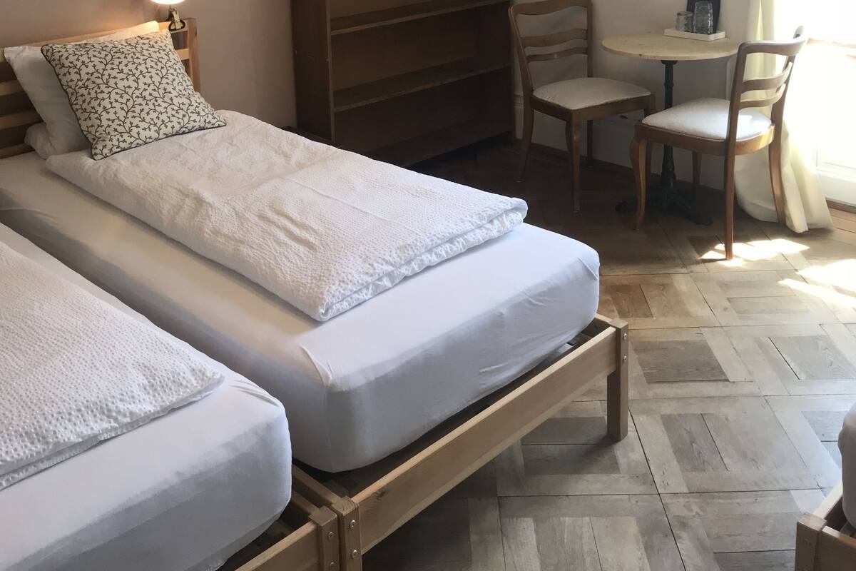 A room with two beds, a wooden nightstand, a small table with chairs, and a bookshelf.