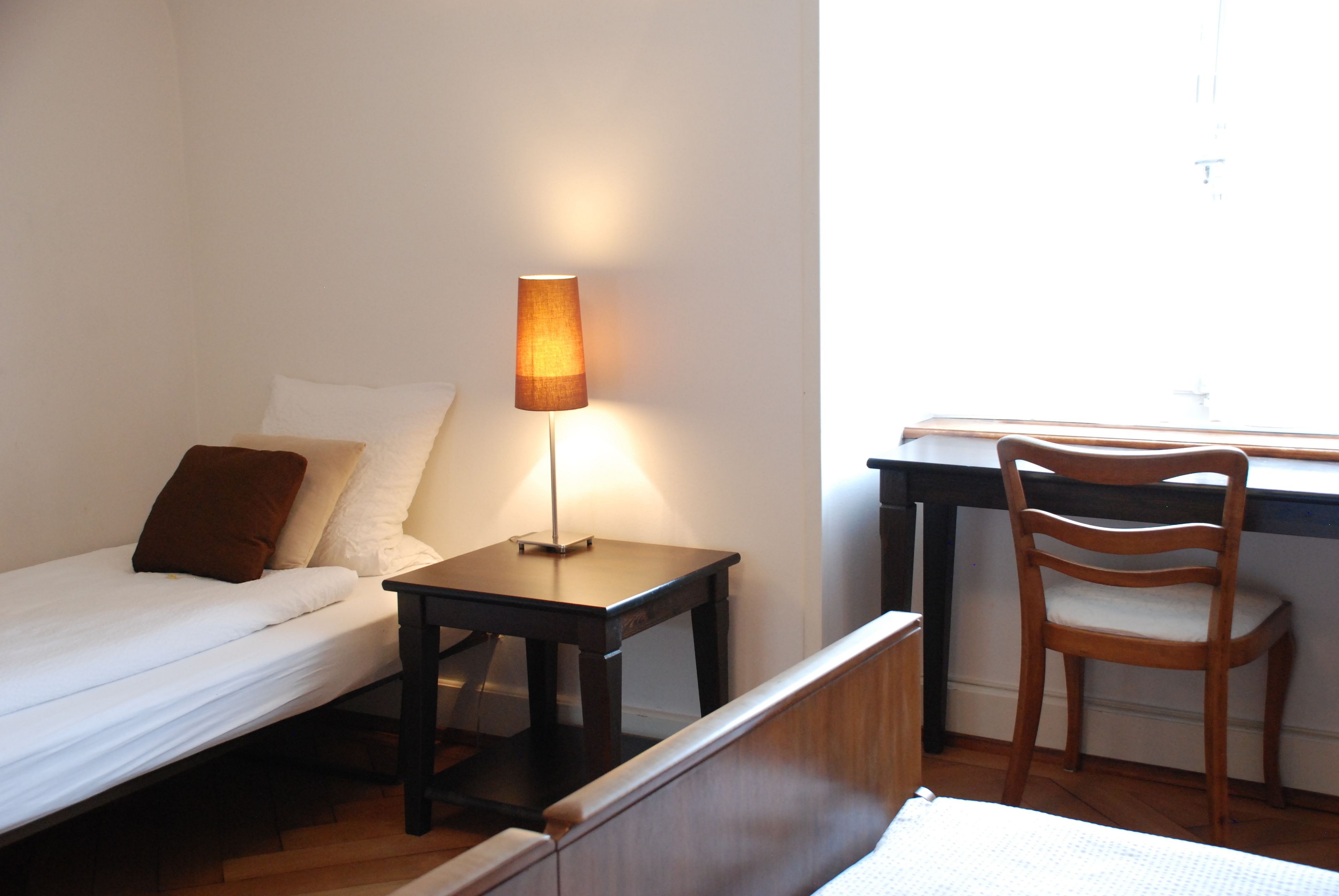 Quarto com cama, mesa de cabeceira com abajur, cadeira de madeira e escrivaninha com cadeira.