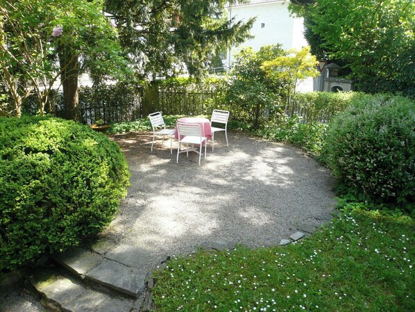 A circular gravel patio with white metal chairs and a table, surrounded by lush greenery and a stone pathway. - The Bed and Breakfast (Lucerne)