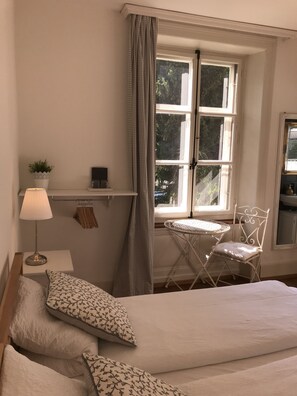 A bedroom with a bed, pillows, a bedside table with a lamp, a small table with a chair, and a window with curtains. - The Bed and Breakfast (Lucerne)