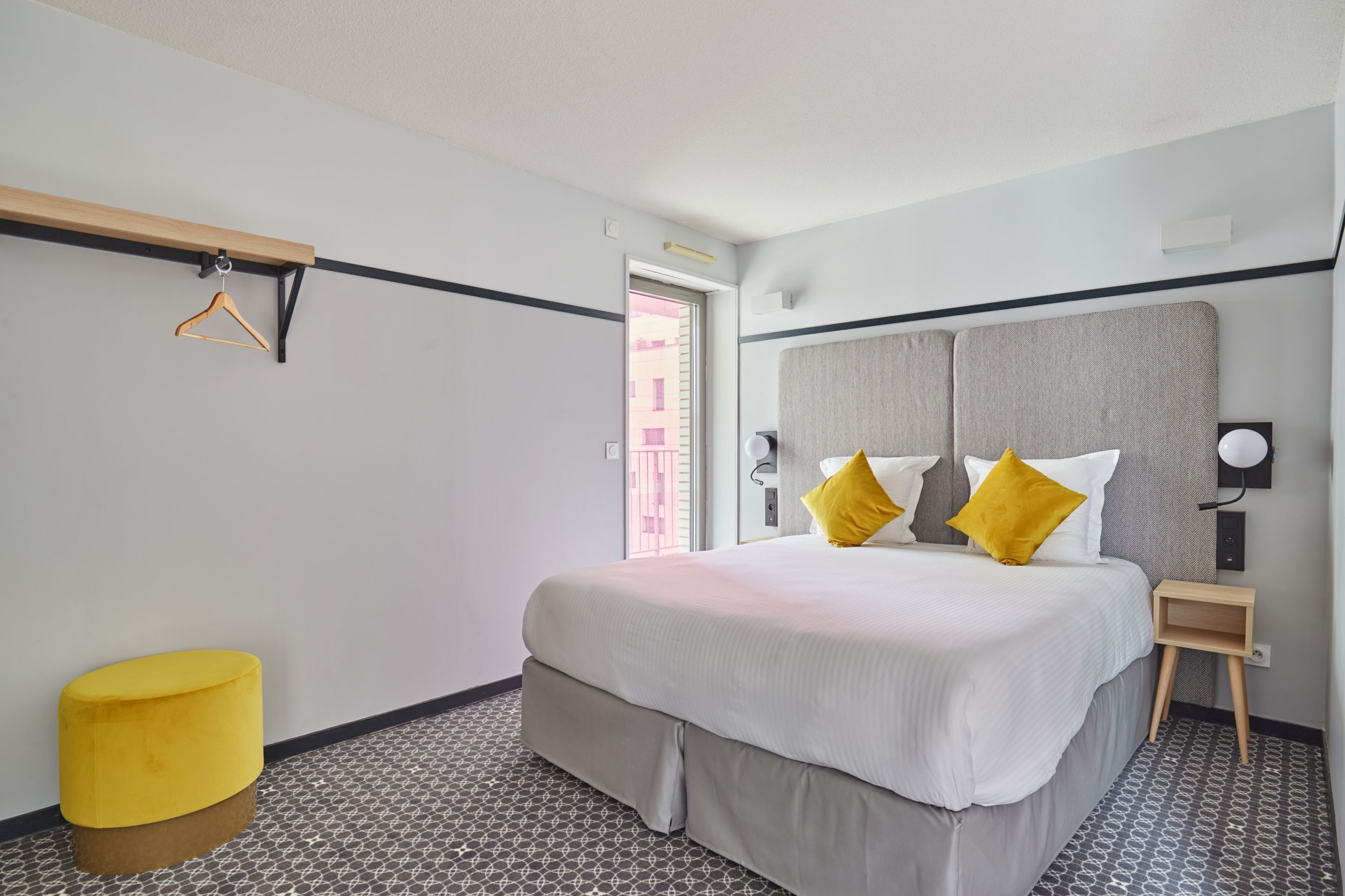 Una habitación de hotel con una cama, una pequeña mesita de noche de madera, un otoman amarillo y una lámpara de pared.