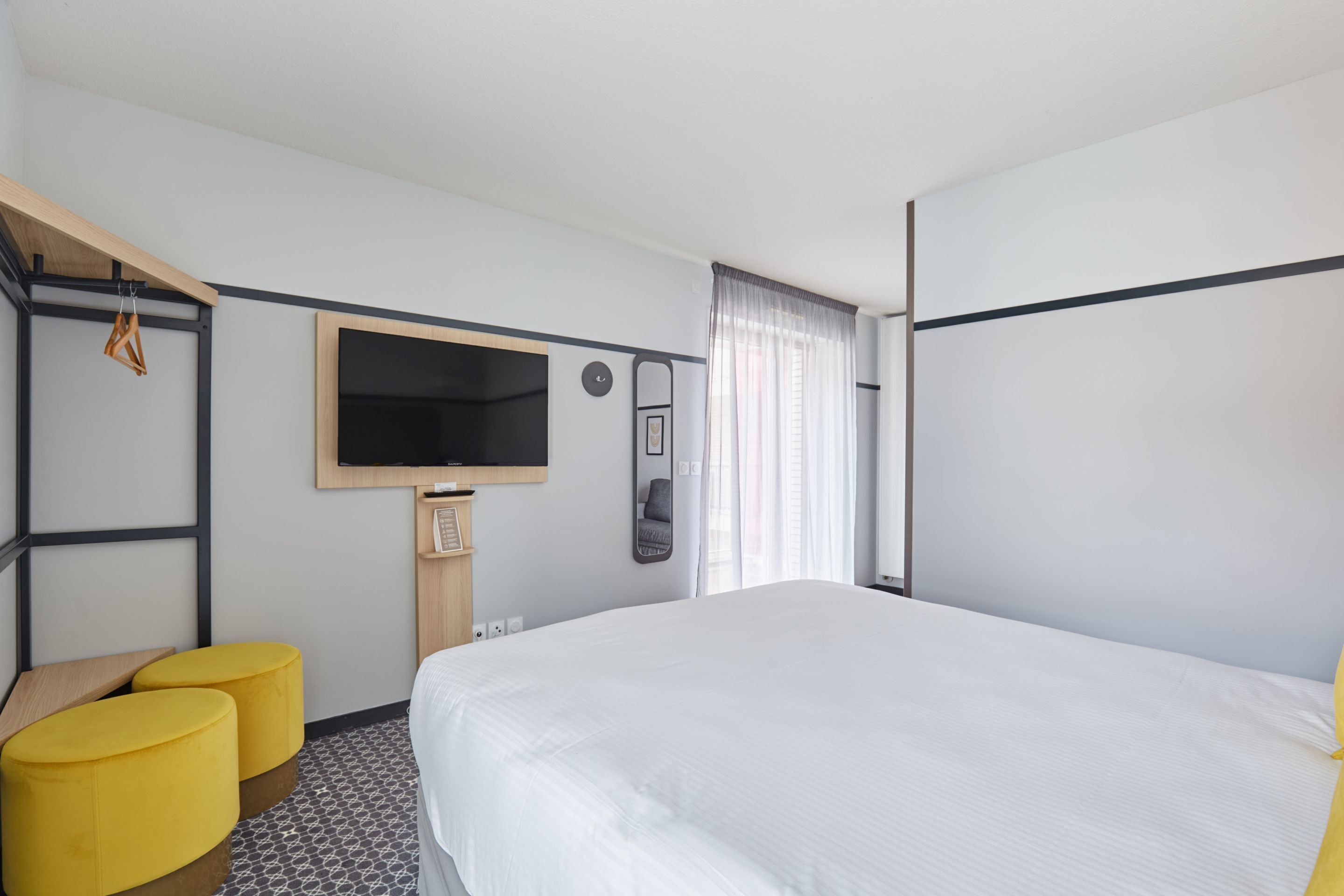 A hotel room with a bed, a TV, a mirror, a clock, and two yellow ottomans.