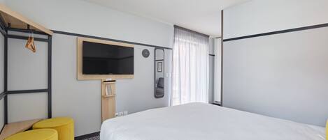 A hotel room with a bed, a TV, a mirror, a clock, and two yellow ottomans.