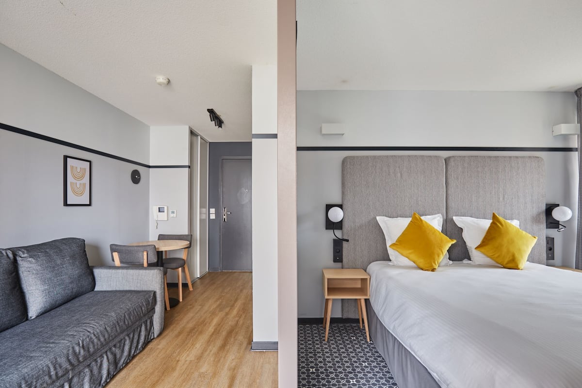 Une chambre d’hôtel avec un canapé gris, une petite table en bois, un lit avec une literie blanche et deux oreillers jaunes, et une tête de lit grise.