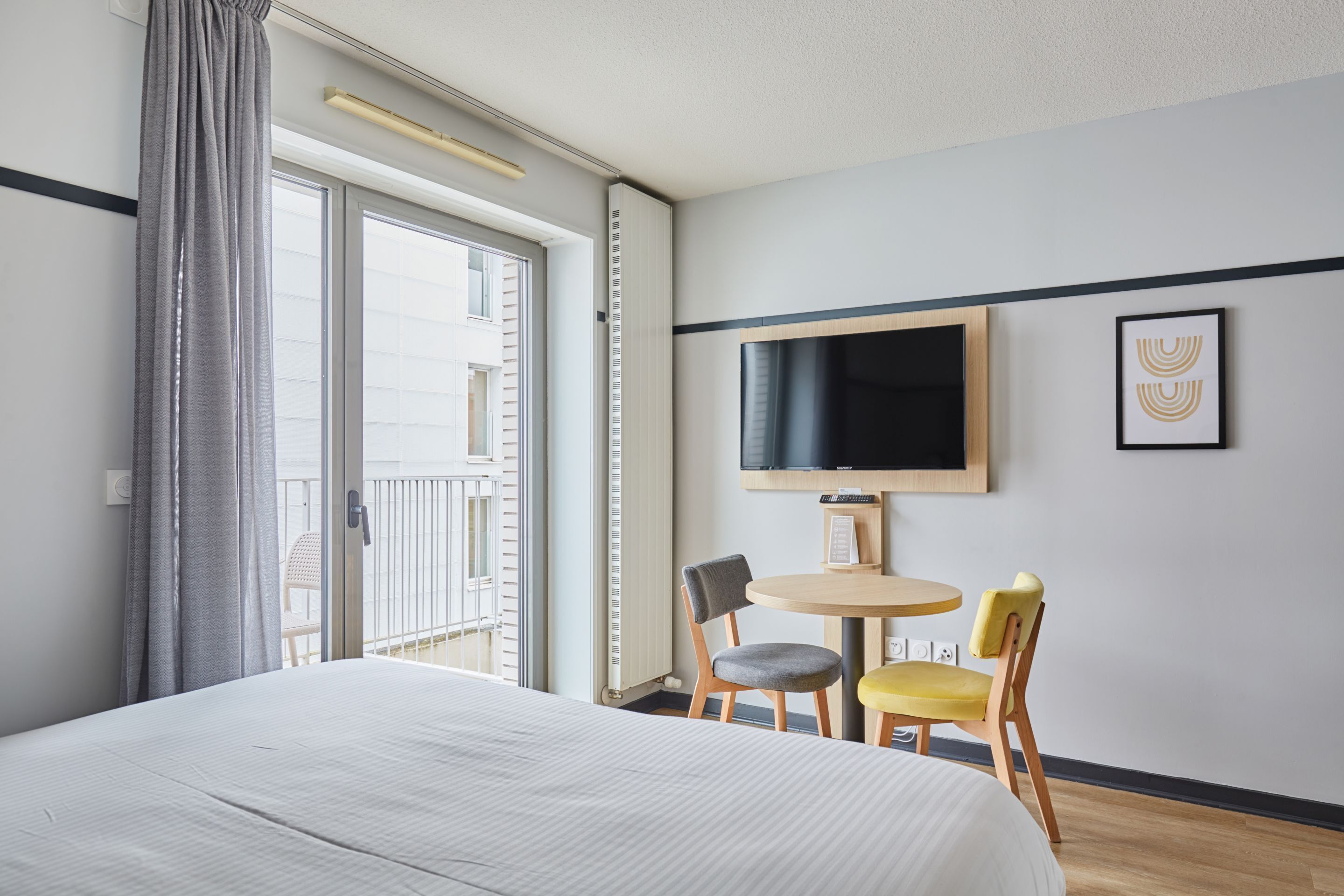 Una camera d'albergo con un letto, una TV fissata al muro, un tavolino rotondo con due sedie e un balcone con vista.