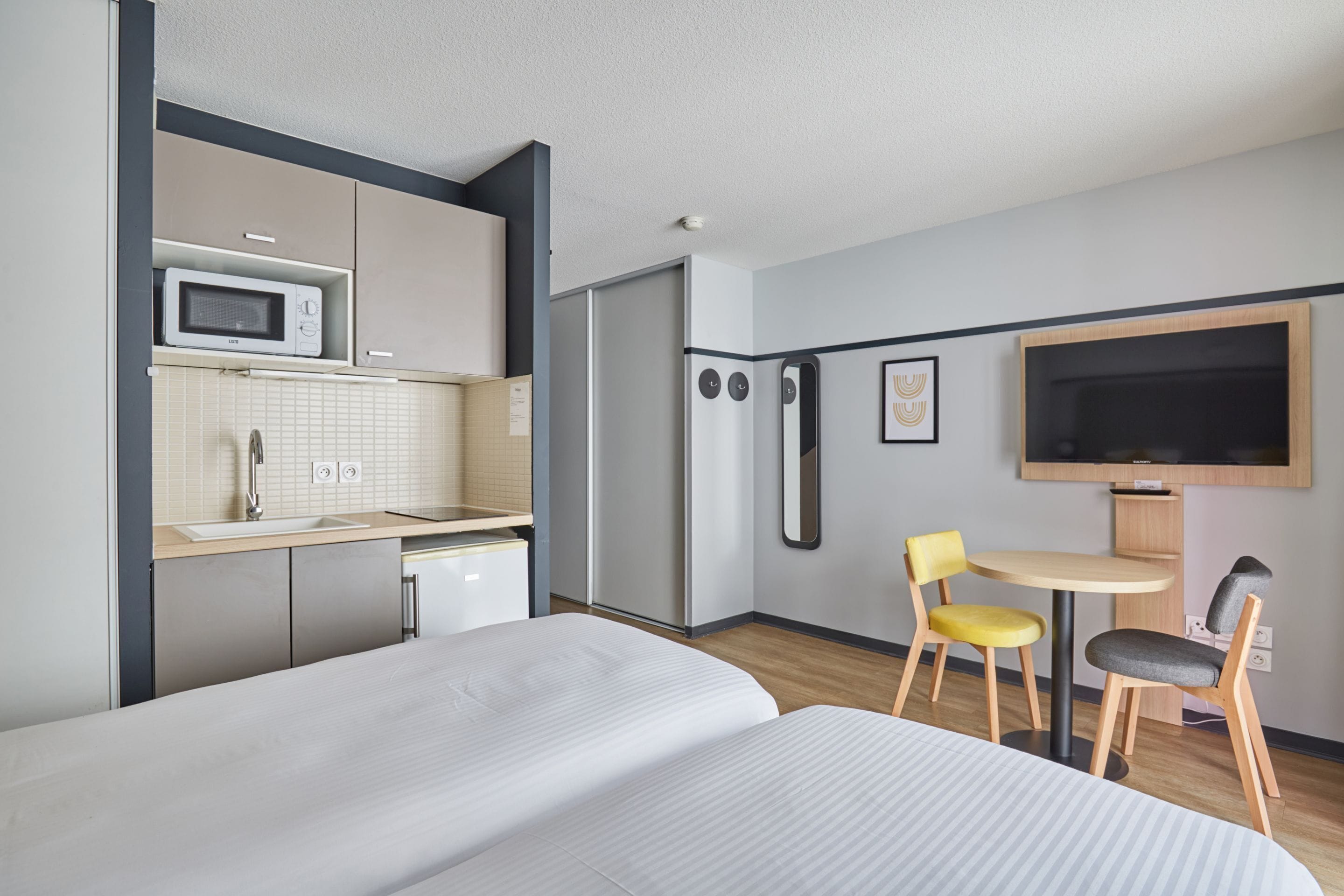 A compact hotel room with a kitchenette, a bed, a small dining table with chairs, and a TV mounted on the wall.