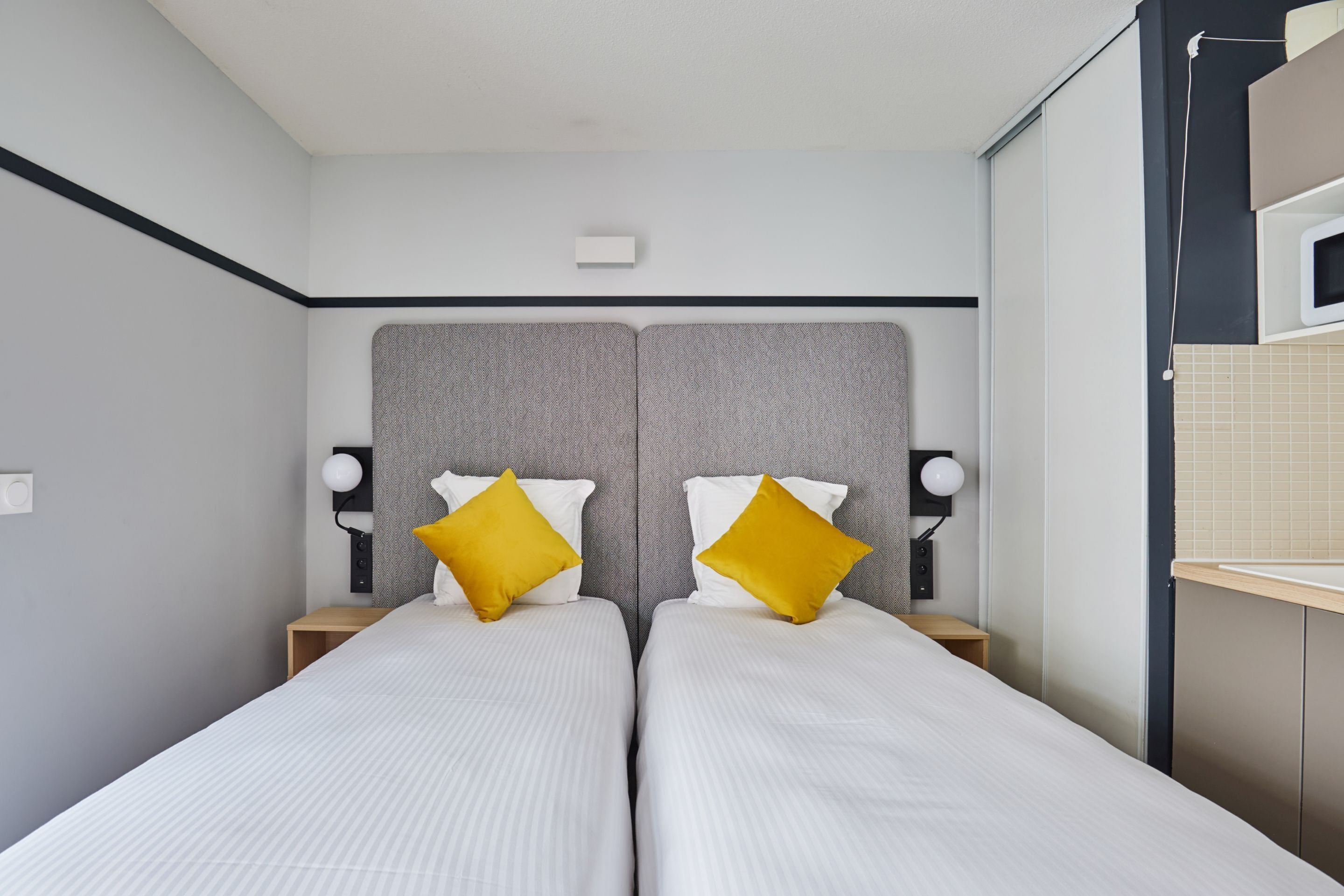 A modern hotel room with two beds, a grey headboard, and a small bedside table with a lamp.