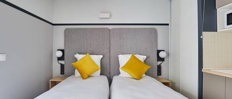 A modern hotel room with two beds, a grey headboard, and a small bedside table with a lamp.