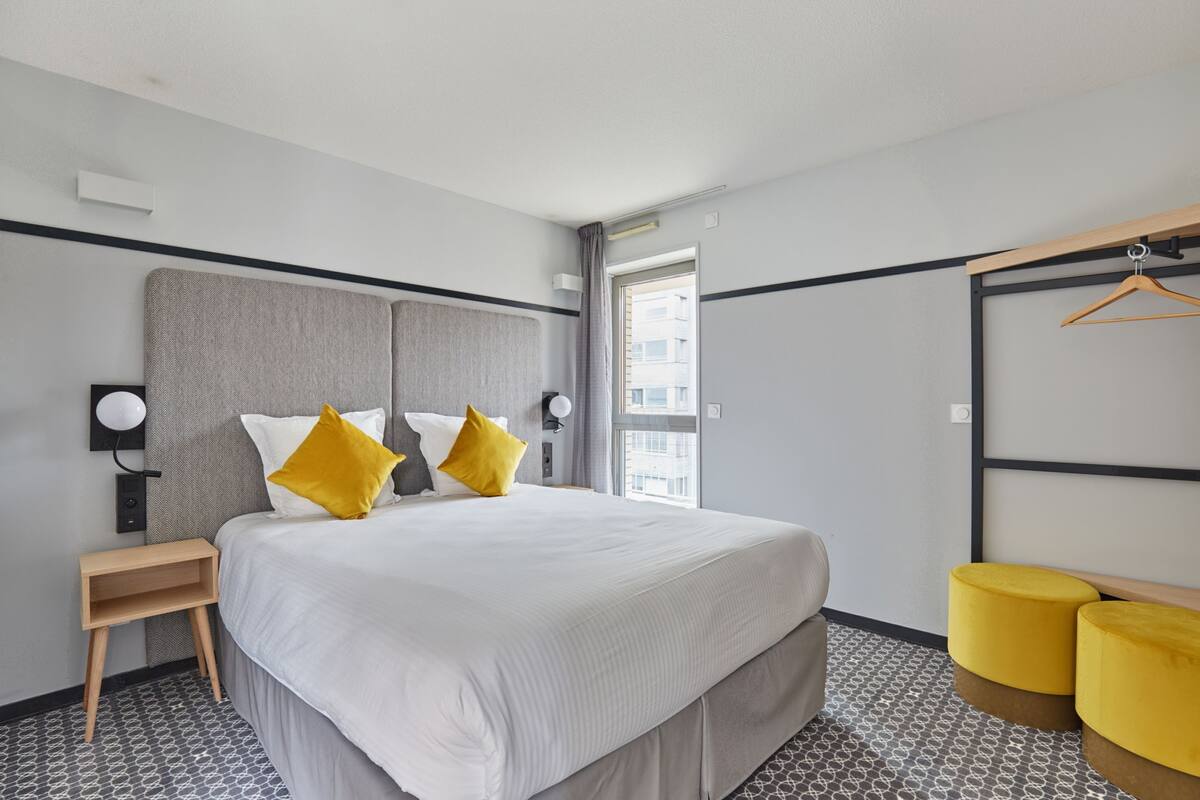 Une chambre d’hôtel moderne, dotée d’un grand lit, de deux tables de chevet, d’un miroir et d’une fenêtre donnant sur des immeubles.