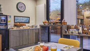 Frühstücksangebot mit Croissants, einem Apfel und Orangensaft auf einem Tablett, eine Uhr an der Wand und Blick durch große Fenster nach draußen.