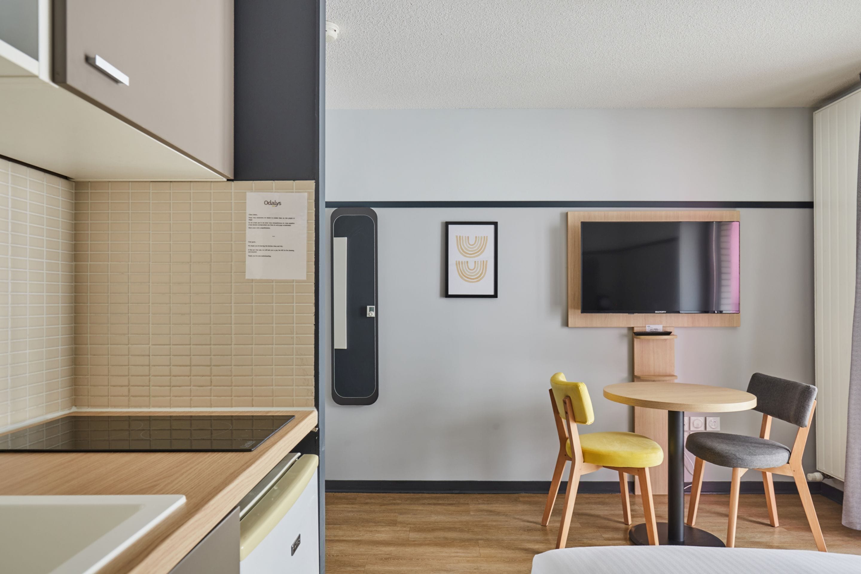 A modern hotel room with a kitchenette, a dining area with two chairs, a flat-screen TV mounted on the wall, and a small table.