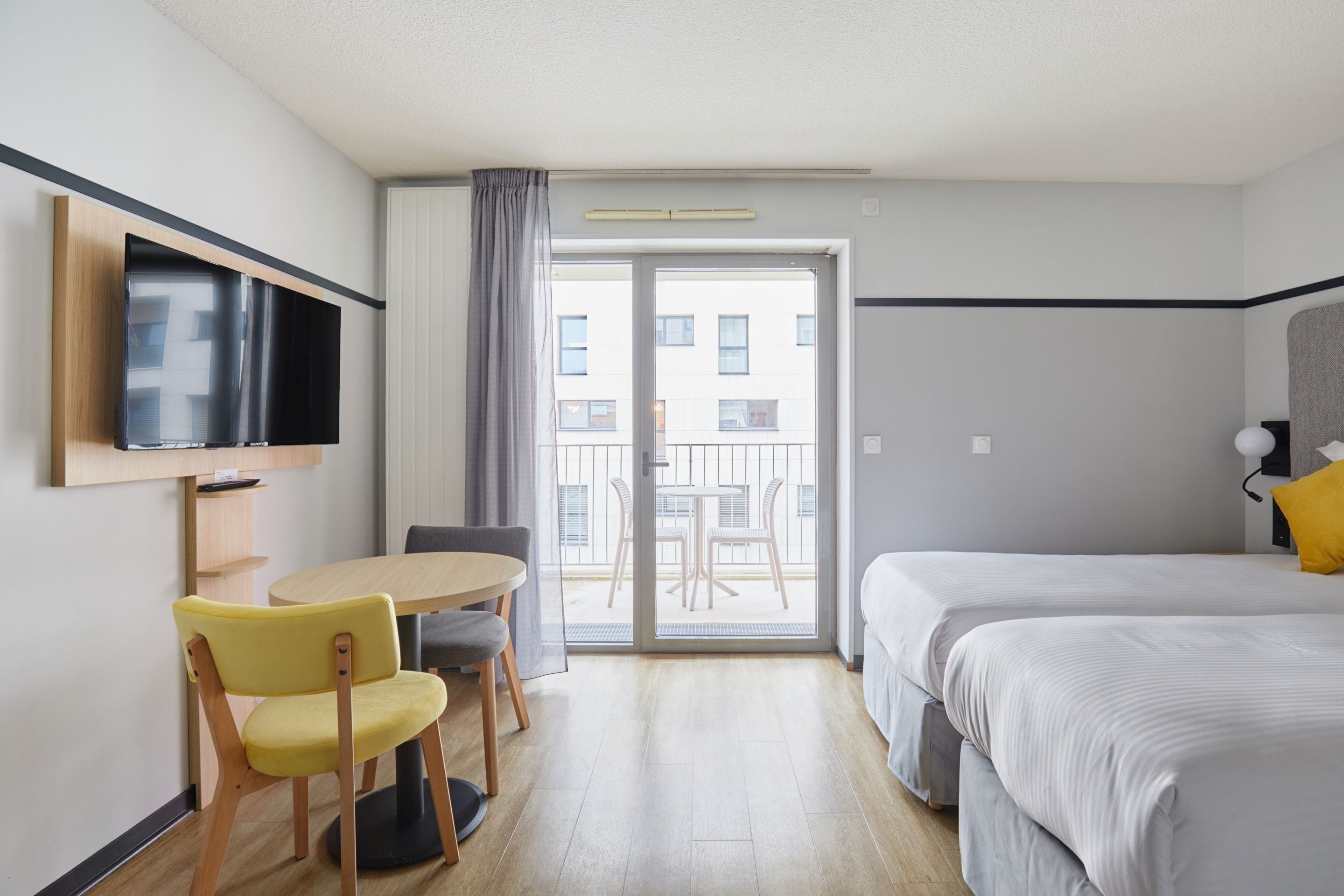 Une chambre d’hôtel comprenant un lit, un téléviseur fixé au mur, un coin repas avec une table et des chaises, et un balcon offrant une vue sur les bâtiments.