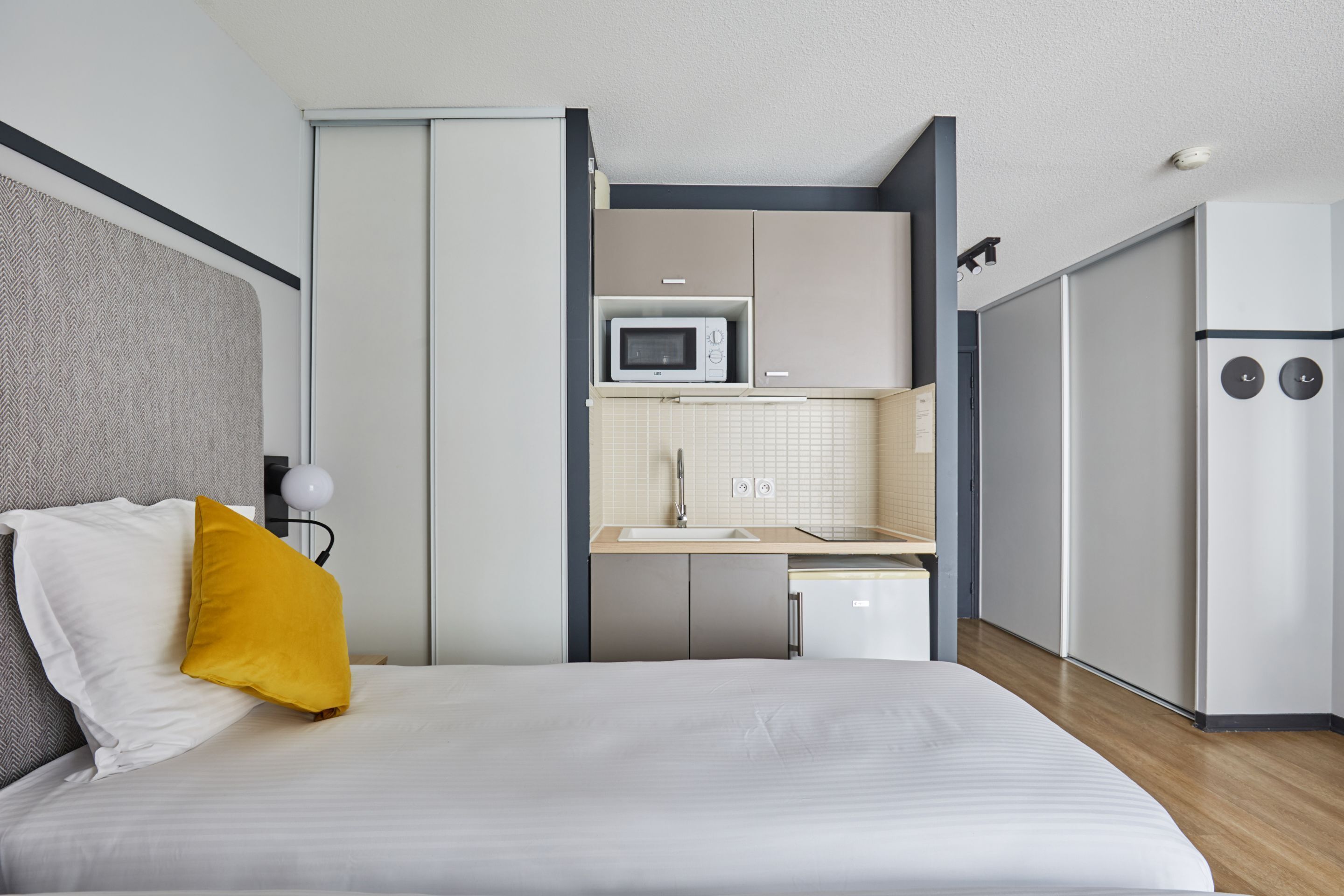 A modern hotel room with a bed, a small kitchenette, and a separate bathroom area.