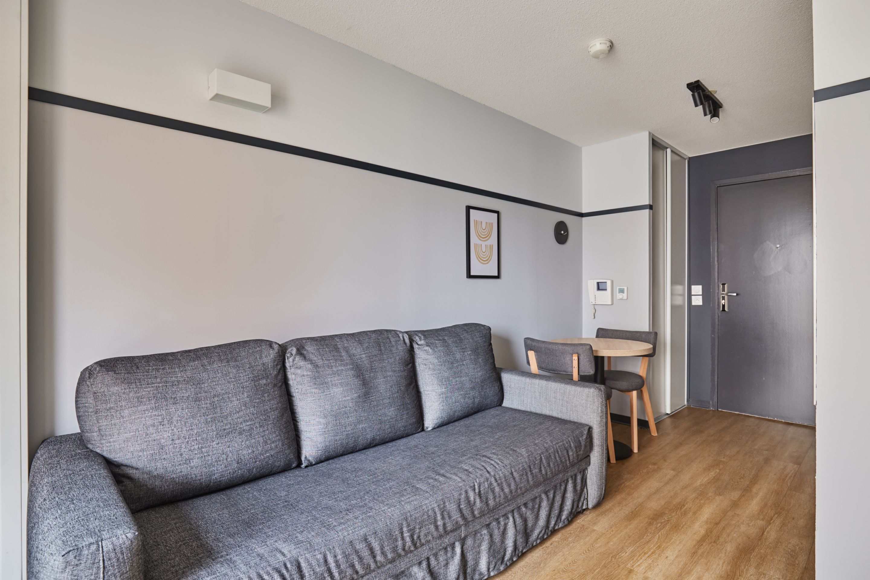 A modern living room with a grey sofa, a small round table, and a chair. There is a door and a wall-mounted light fixture.