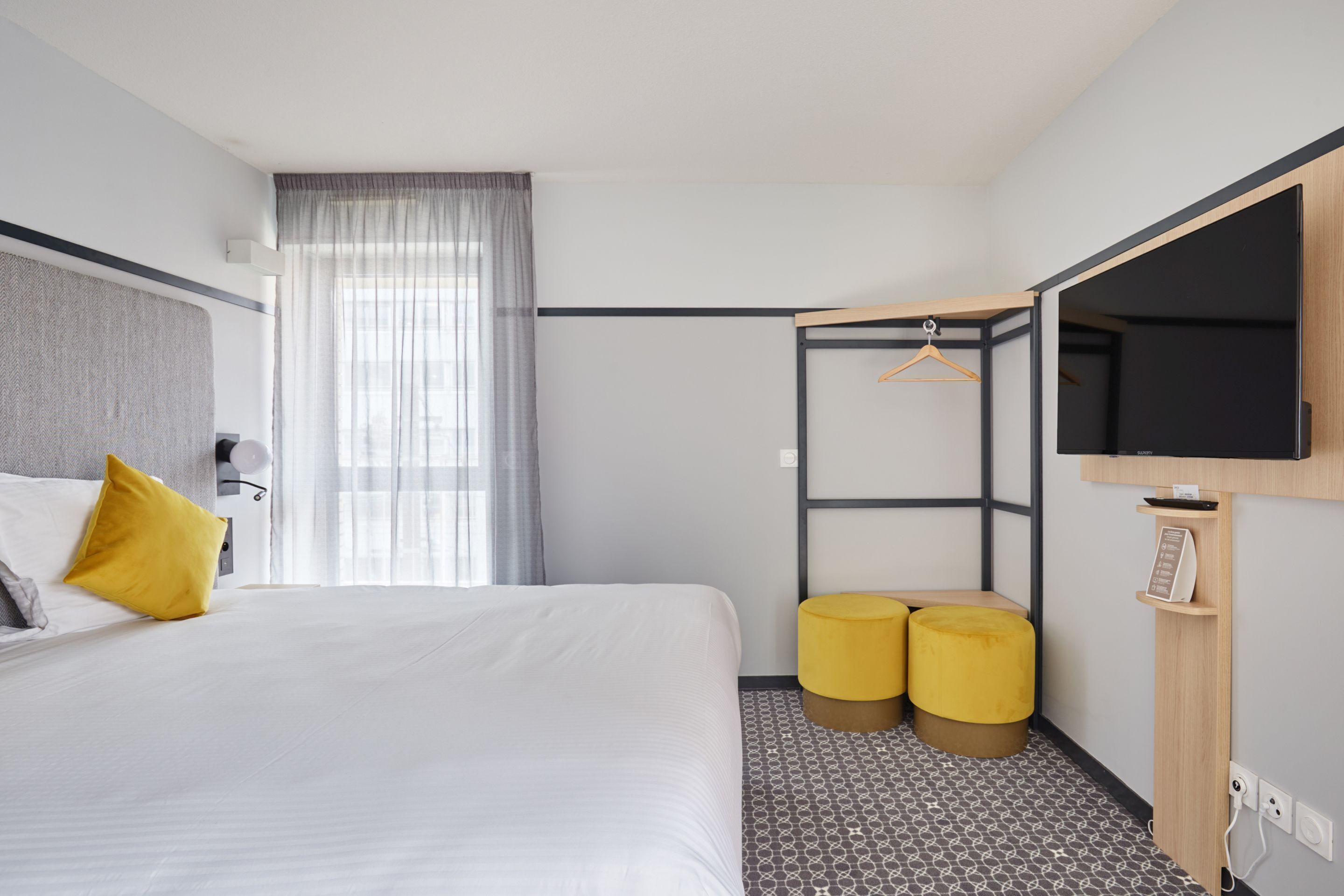 A hotel room with a bed, a TV mounted on the wall, a small seating area with two ottomans, and a window with curtains.