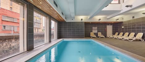 An indoor swimming pool with clear glass walls, a tiled floor, and lounge chairs along the side.