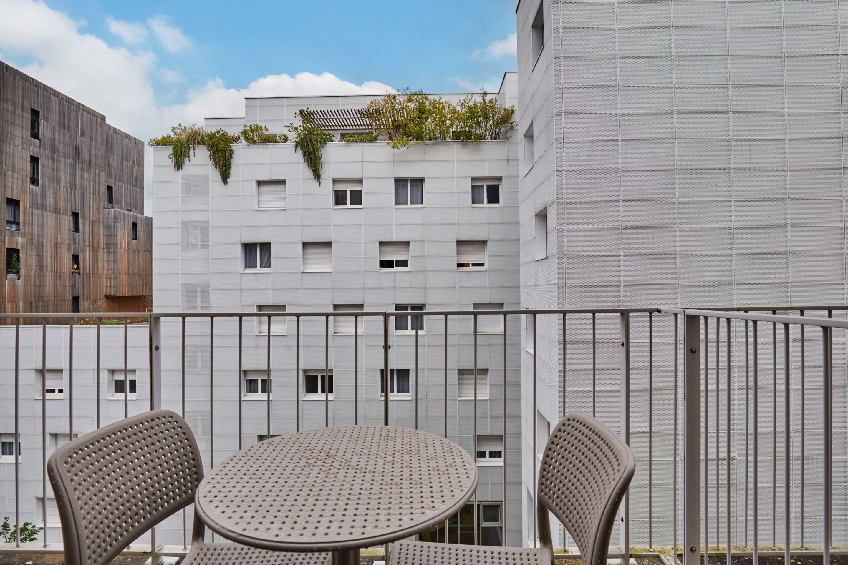 Un balcón con una mesa redonda y dos sillas, con vistas a un edificio de múltiples ventanas y fachada gris.