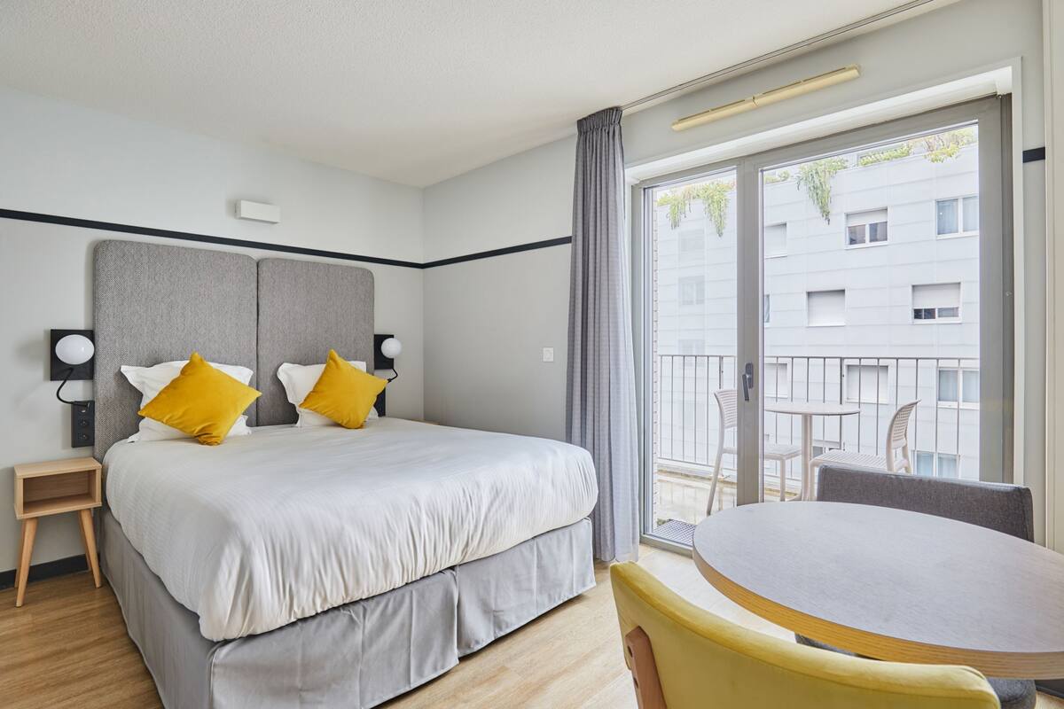 Une chambre d’hôtel comprenant un lit, des tables de chevet, une petite table et un balcon offrant une vue sur les bâtiments et la verdure.