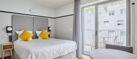 A hotel room with a bed, bedside tables, a small table, and a balcony with a view of buildings and greenery.