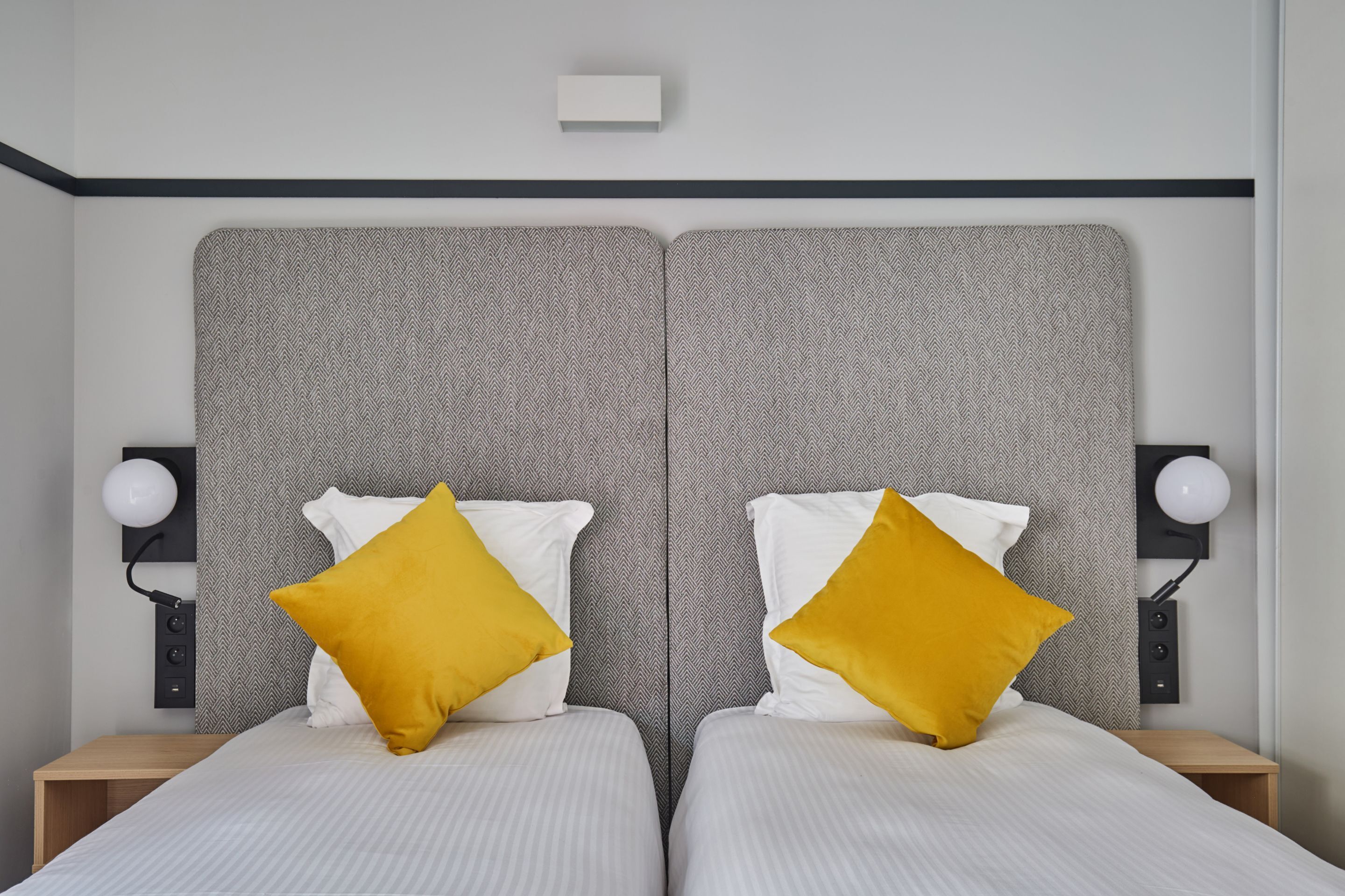 A double bed with two pillows, a nightstand, a wall-mounted lamp, and a small shelf with a light fixture.